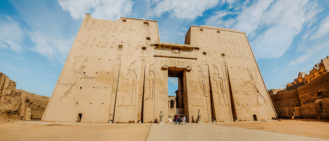 Tourists admire the towering ancient Edfu Temple entrance with detailed carved reliefs under a bright sky.