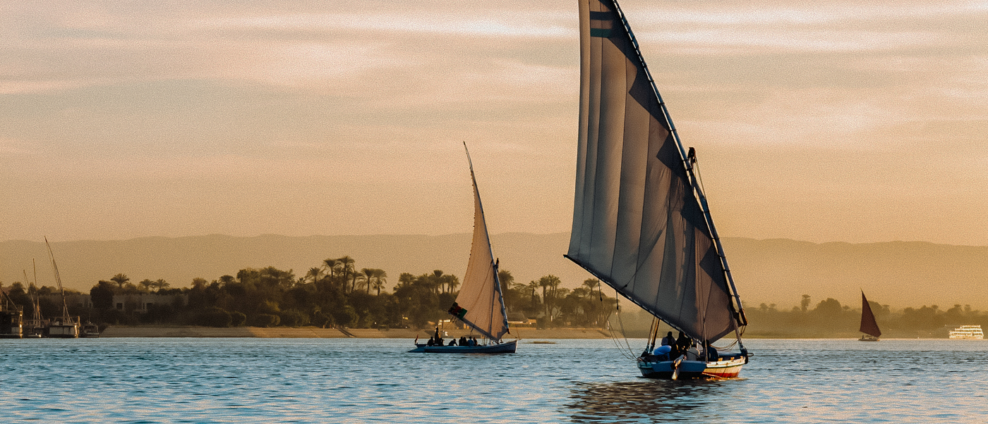 Two feluccas sail peacefully on the Nile River near Luxor, with a golden sky and green banks.