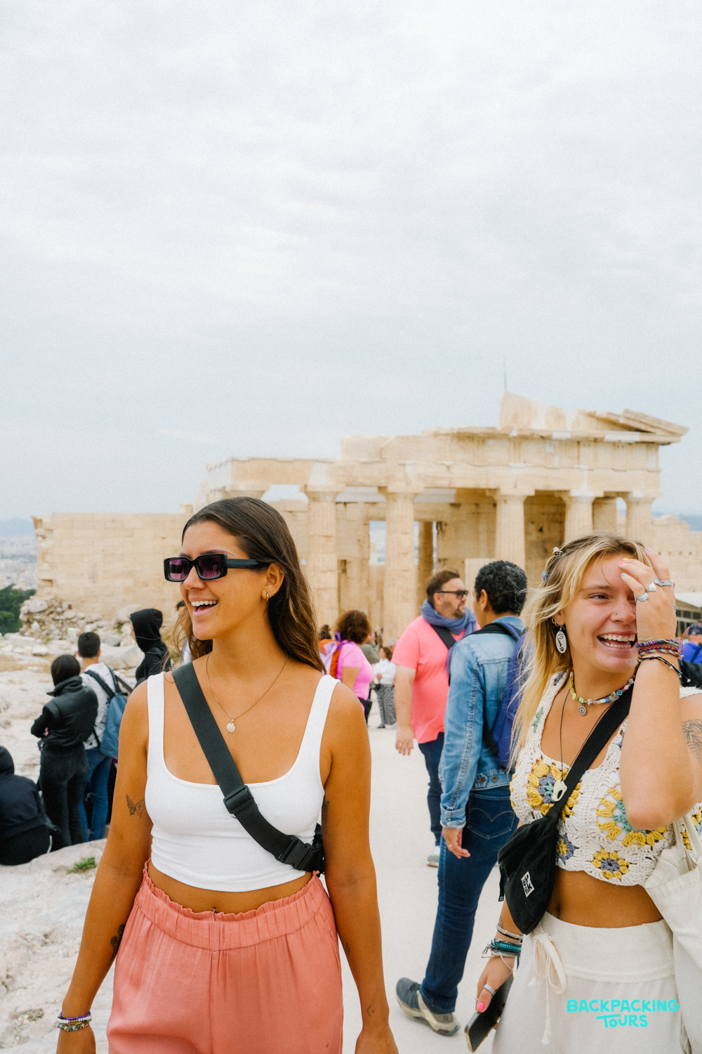 Two happy women laughing while exploring the ancient Acropolis in Athens with other tourists.
