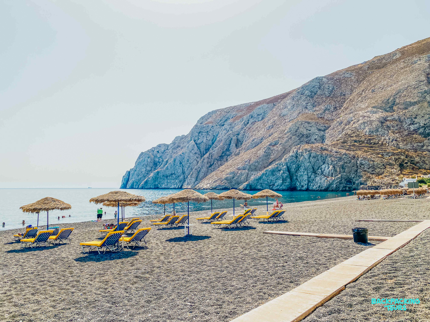 A tranquil dark sand beach in Mykonos, with sun loungers and straw umbrellas by the sea.