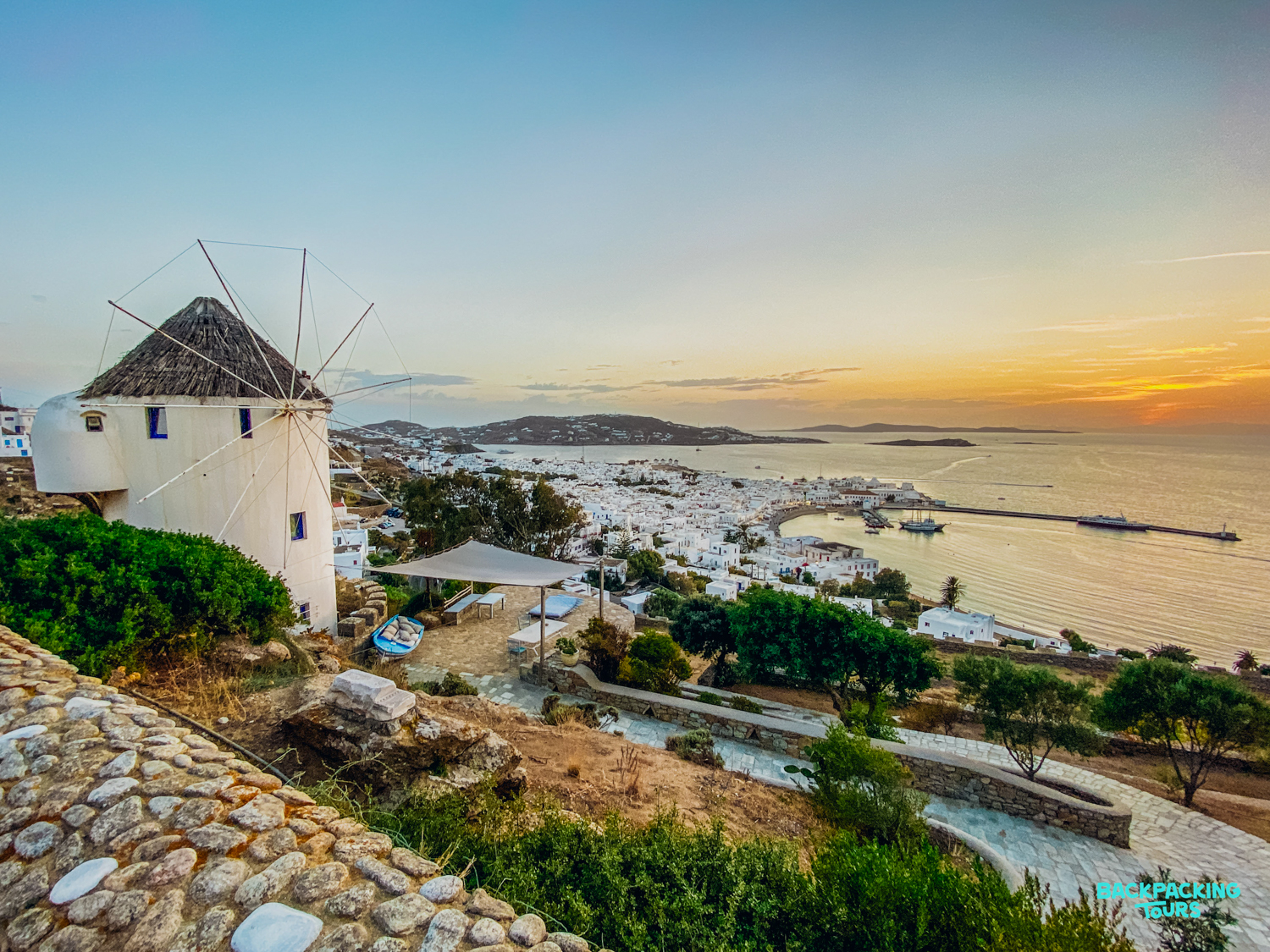 A classic Mykonos windmill overlooks the whitewashed town and tranquil bay under a beautiful sunset.