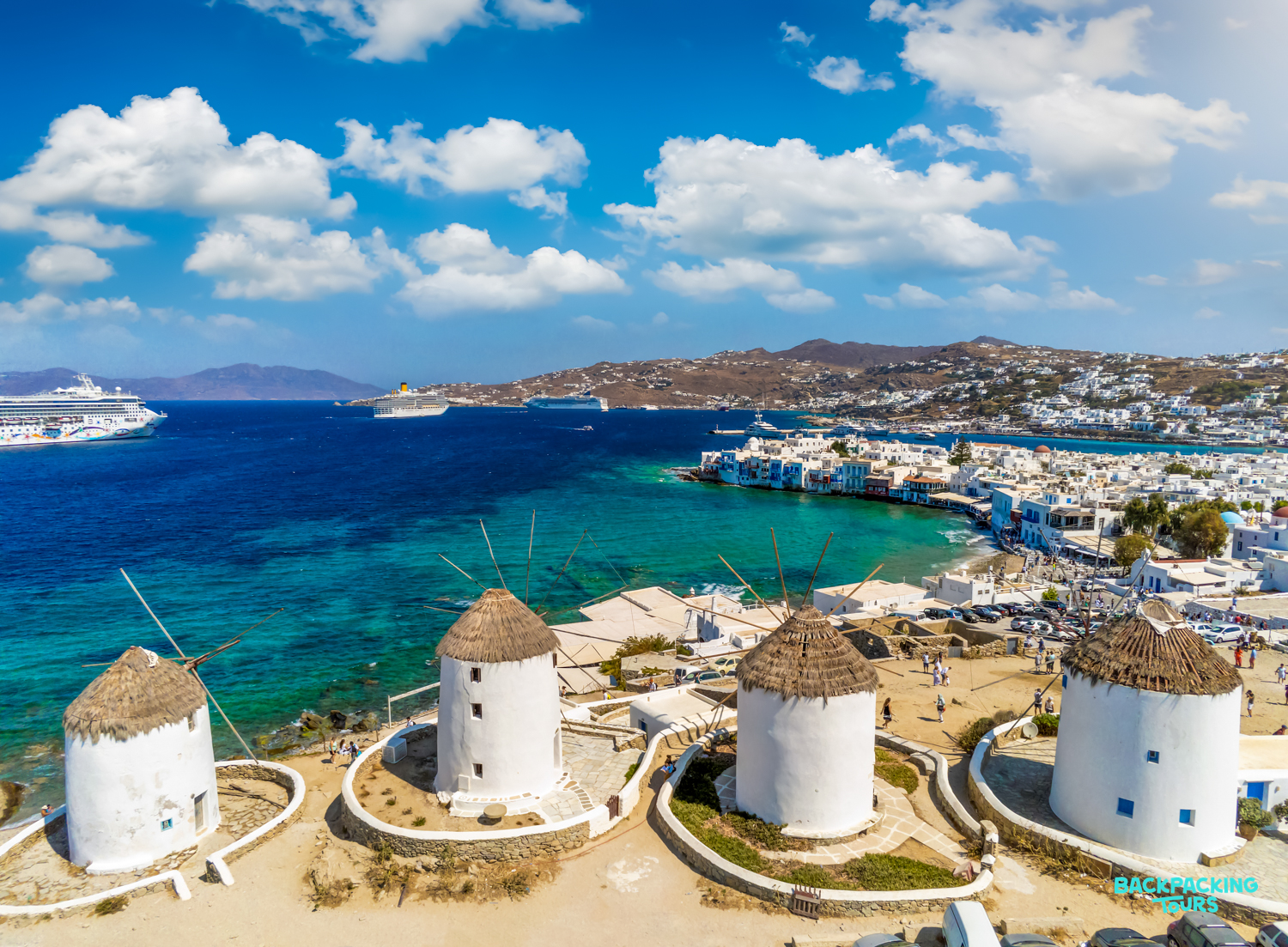 Four iconic windmills stand proudly overlooking the vibrant blue Aegean Sea and whitewashed Mykonos town.