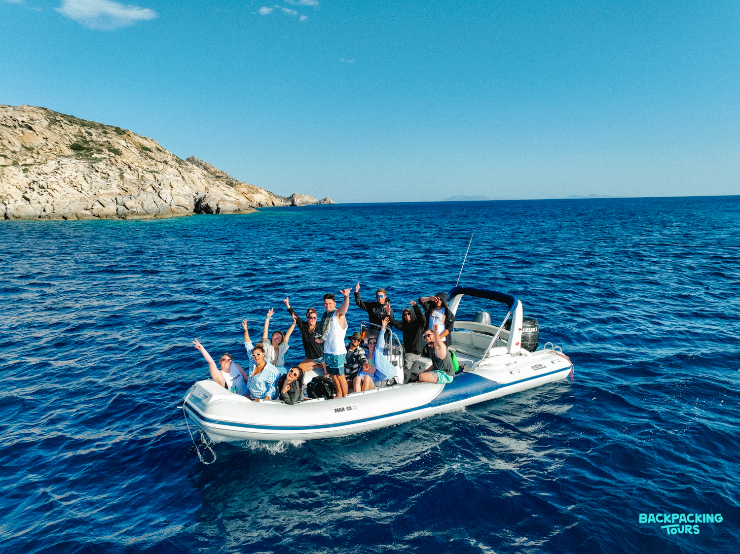 A group of happy backpackers waving from a speedboat on the sparkling blue waters off Ios, Greece.