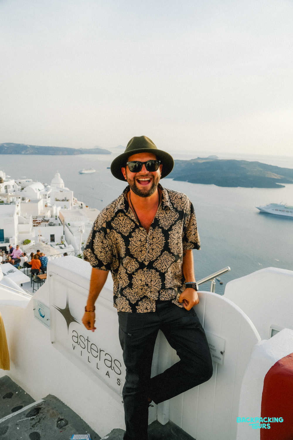 A happy man in a hat and sunglasses smiles, overlooking the white buildings and blue sea of Santorini.