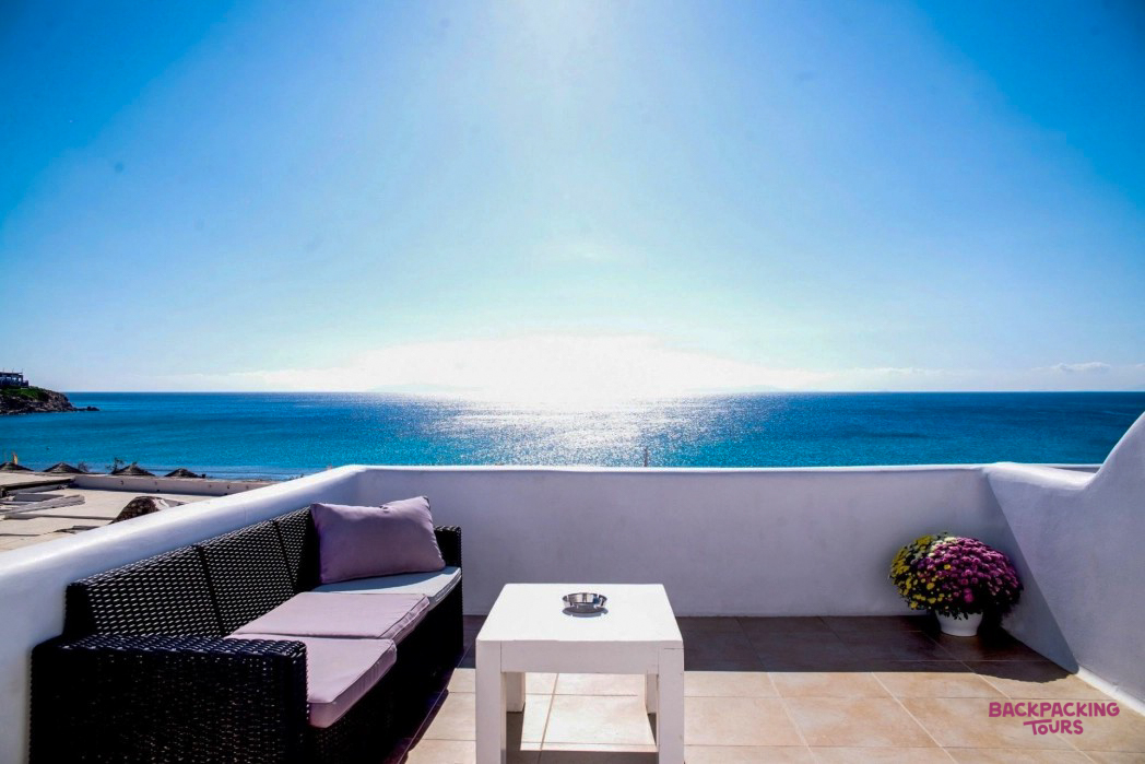 Bright Mykonos balcony with a comfortable sofa and coffee table overlooking the sparkling blue Aegean Sea.