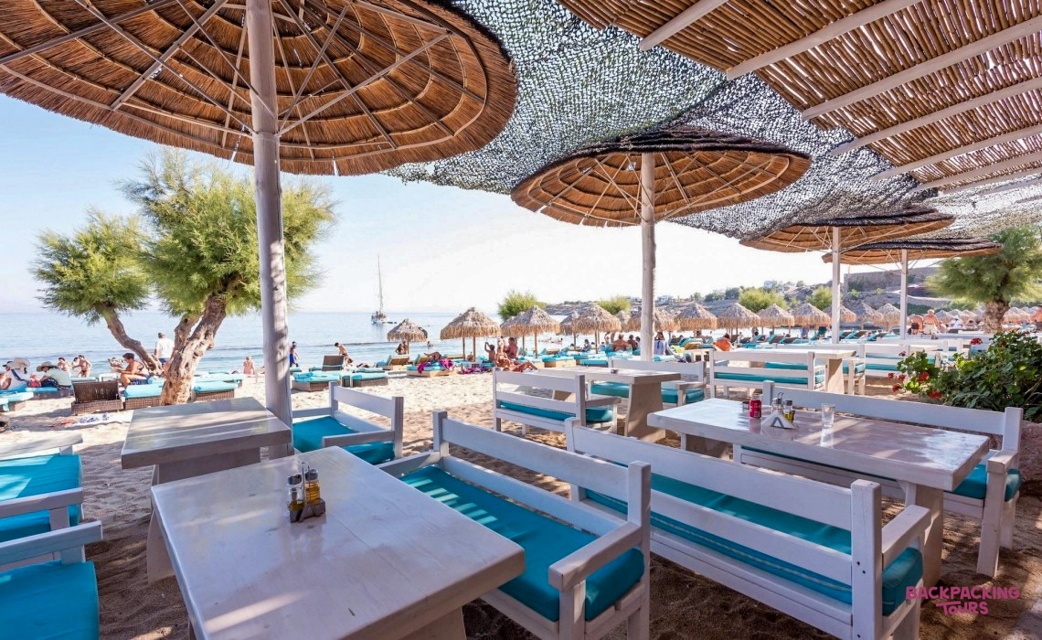 White tables and turquoise seating under straw umbrellas at a sunny beach club in Mykonos.