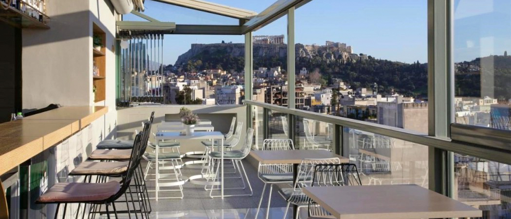 A modern rooftop bar with tables and chairs overlooking the city of Athens and the Acropolis.