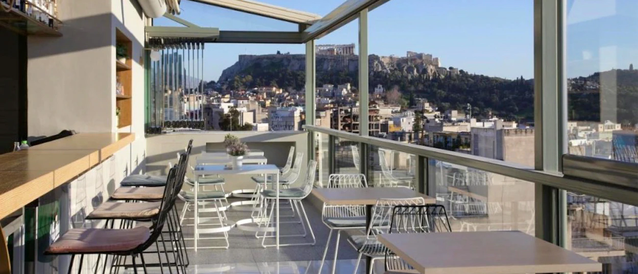 A modern rooftop bar with tables and chairs overlooking the city of Athens and the Acropolis.