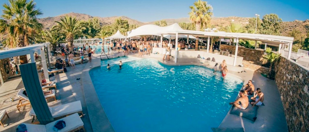 A lively pool party with many people swimming and socializing under palm trees in Ios.