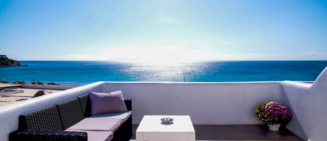 Bright Mykonos terrace with a sofa, coffee table, and flowers, facing the shimmering blue sea.