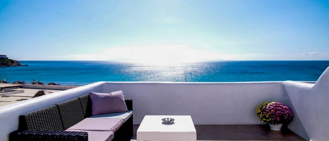 Bright Mykonos terrace with a sofa, coffee table, and flowers, facing the shimmering blue sea.