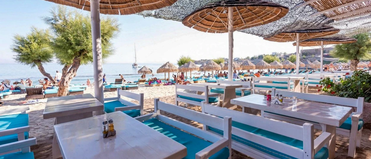 Sunny Mykonos beach club with white tables, blue cushions, and many people relaxing by the sea.