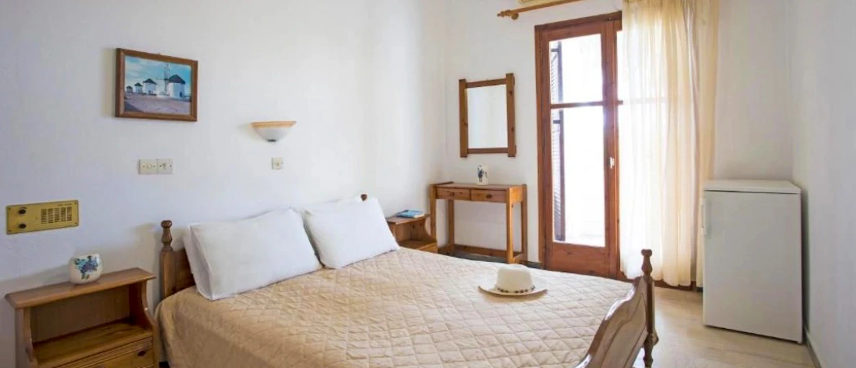 A simple, bright hotel room in Santorini with a double bed and a painting of island architecture.