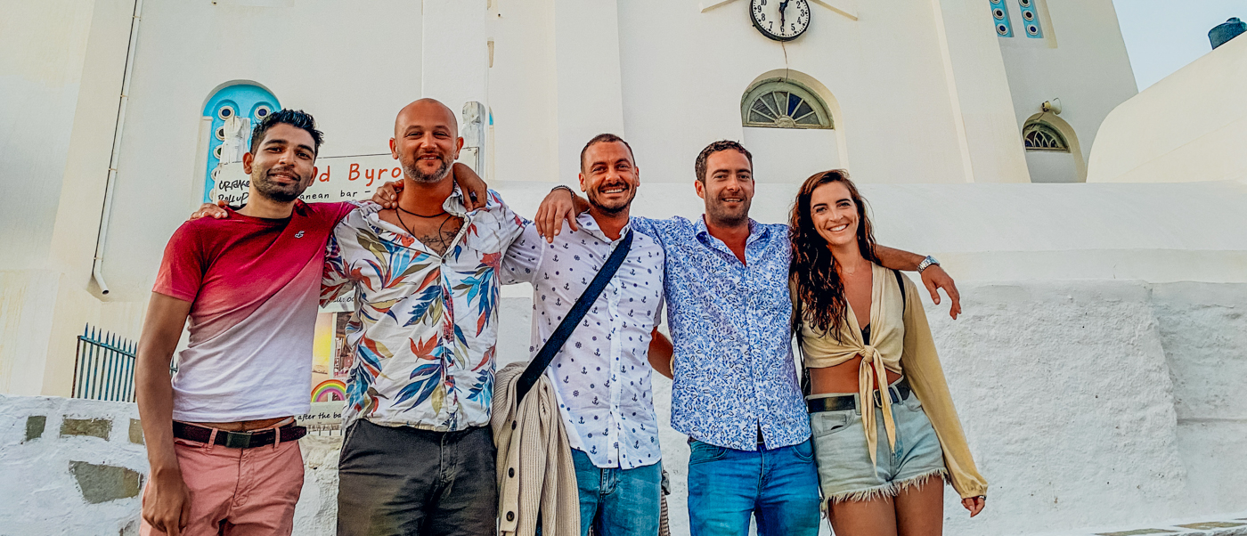 Five happy friends pose arm-in-arm in front of a white Greek building in Ios.