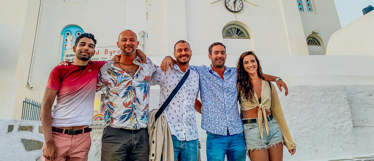 Five happy friends pose arm-in-arm in front of a white Greek building in Ios.