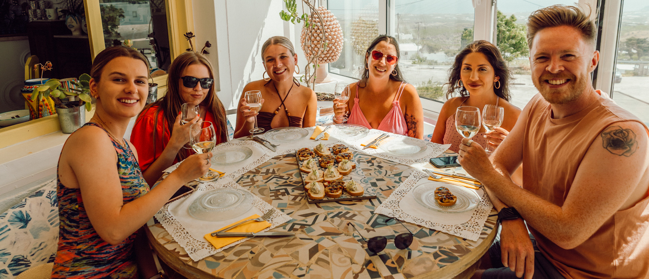 Six happy friends smiling and toasting with wine glasses at a table in Santorini.