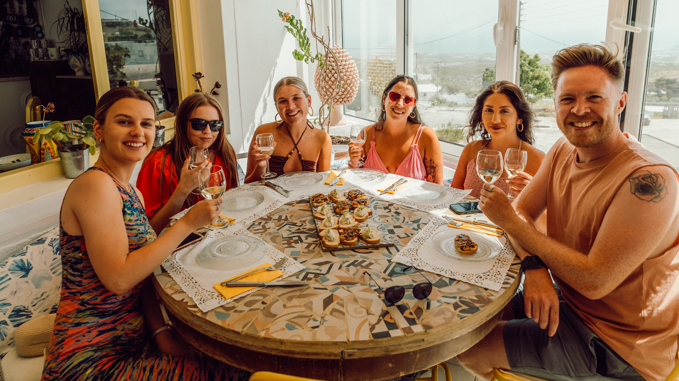 Six happy friends toast with wine around a mosaic table in a bright Santorini room.