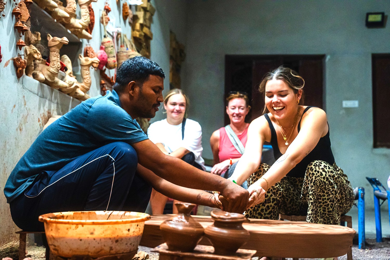 A local potter guides a smiling traveller's hands at the pottery wheel in a Bishnoi village as two others look on.