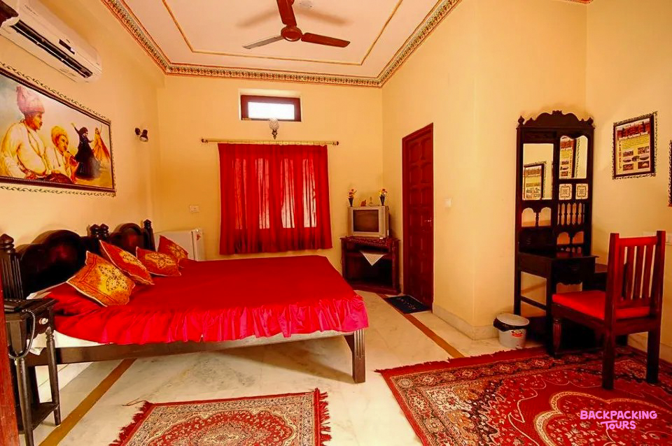 A vibrant haveli room in Jodhpur with a large red bed, ornate rugs, and traditional Indian artwork.
