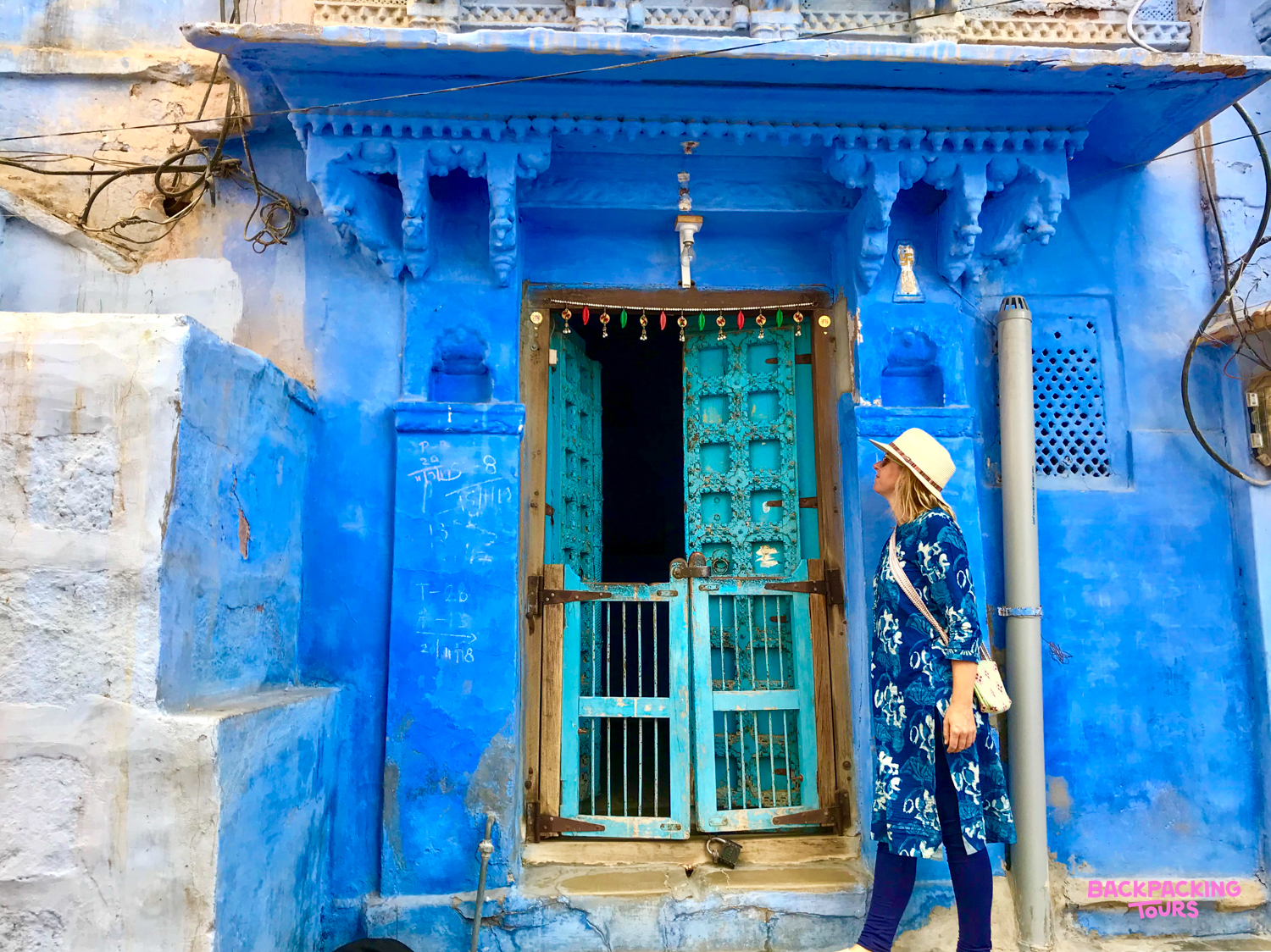 A traveler in a hat stands before a vibrant blue building with an ornate door in Jodhpur.