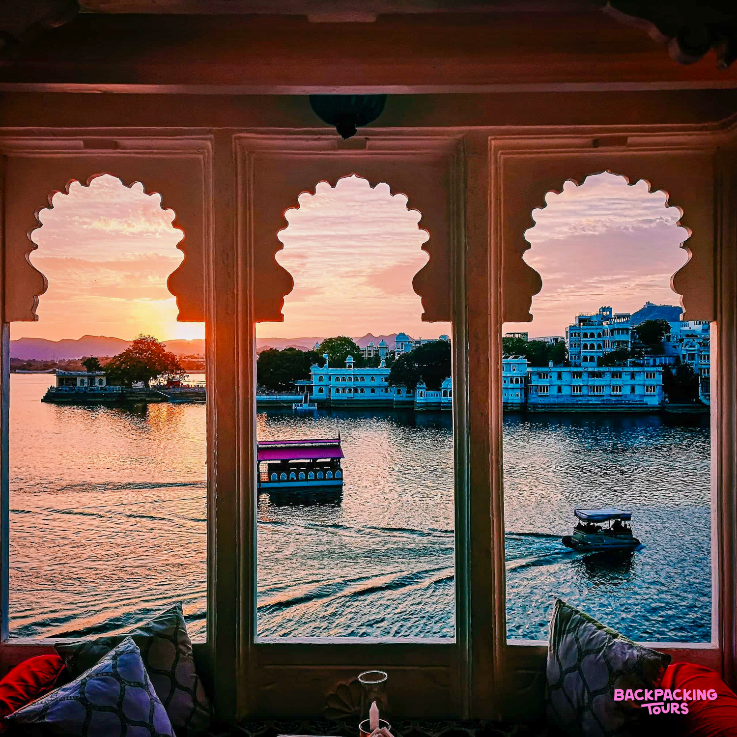 Udaipur's Lake Pichola at sunset, framed by ornate arched windows, with boats and white palaces.
