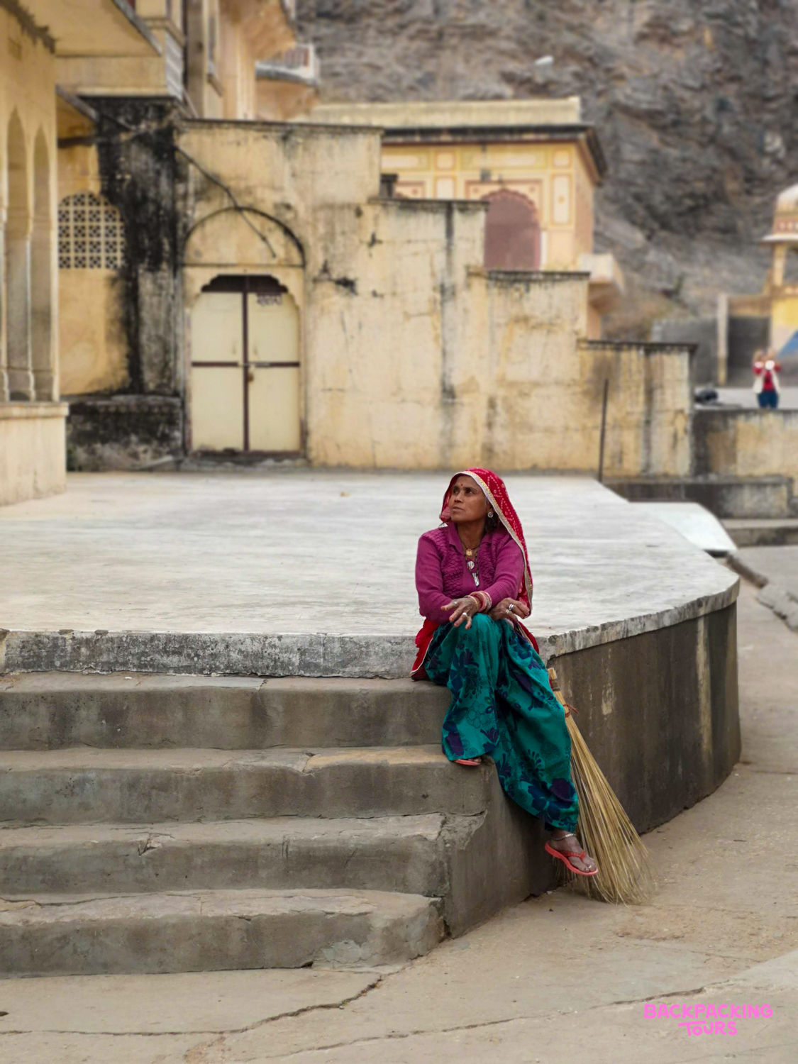 A woman in vibrant traditional dress sits on stone steps in Udaipur, looking thoughtfully upwards.
