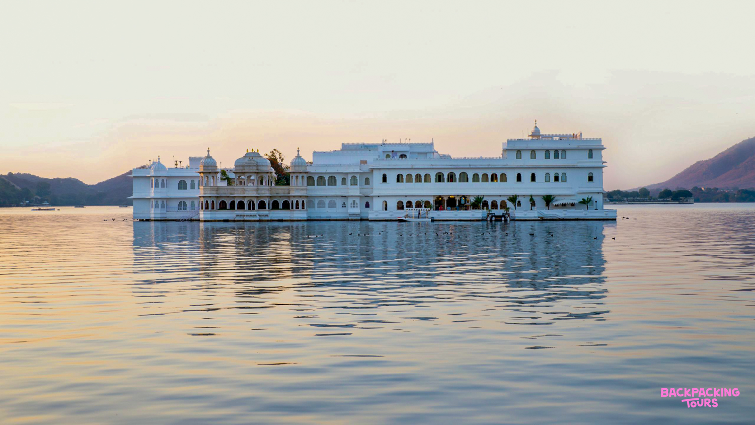 A majestic white palace floats on Lake Pichola in Udaipur, beautifully reflecting the sunset sky.
