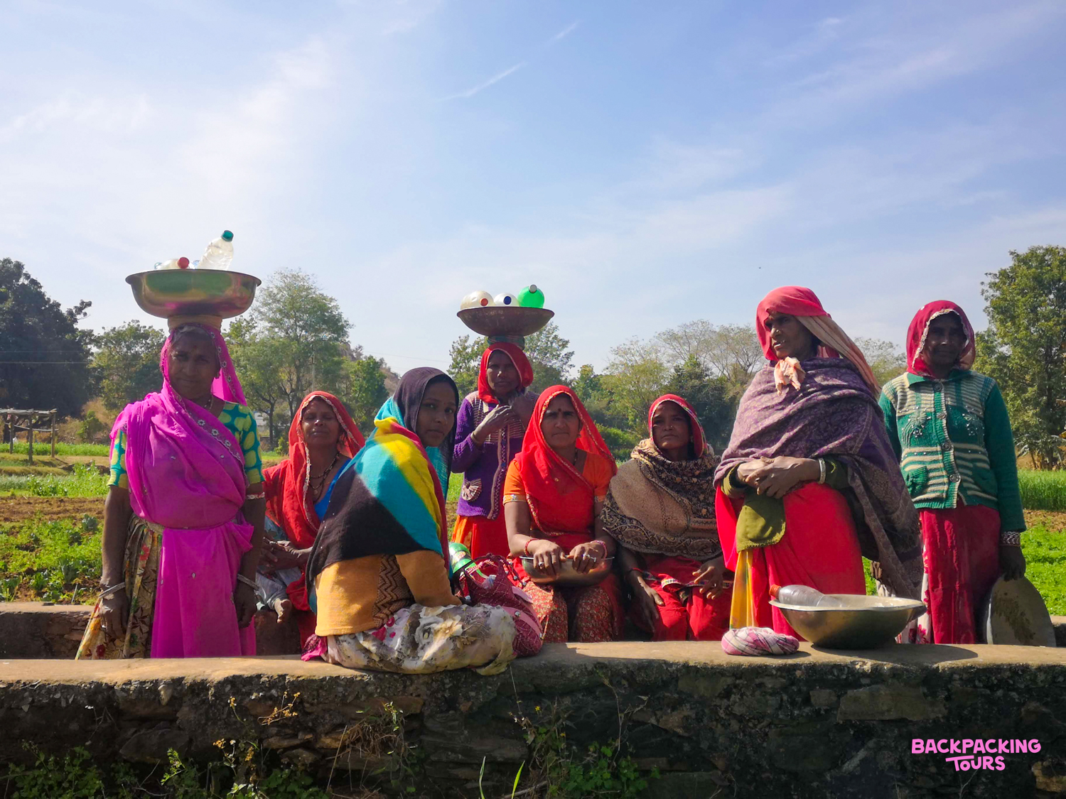 Local women in colorful traditional Indian attire gather by a low wall in a sunny, rural setting.