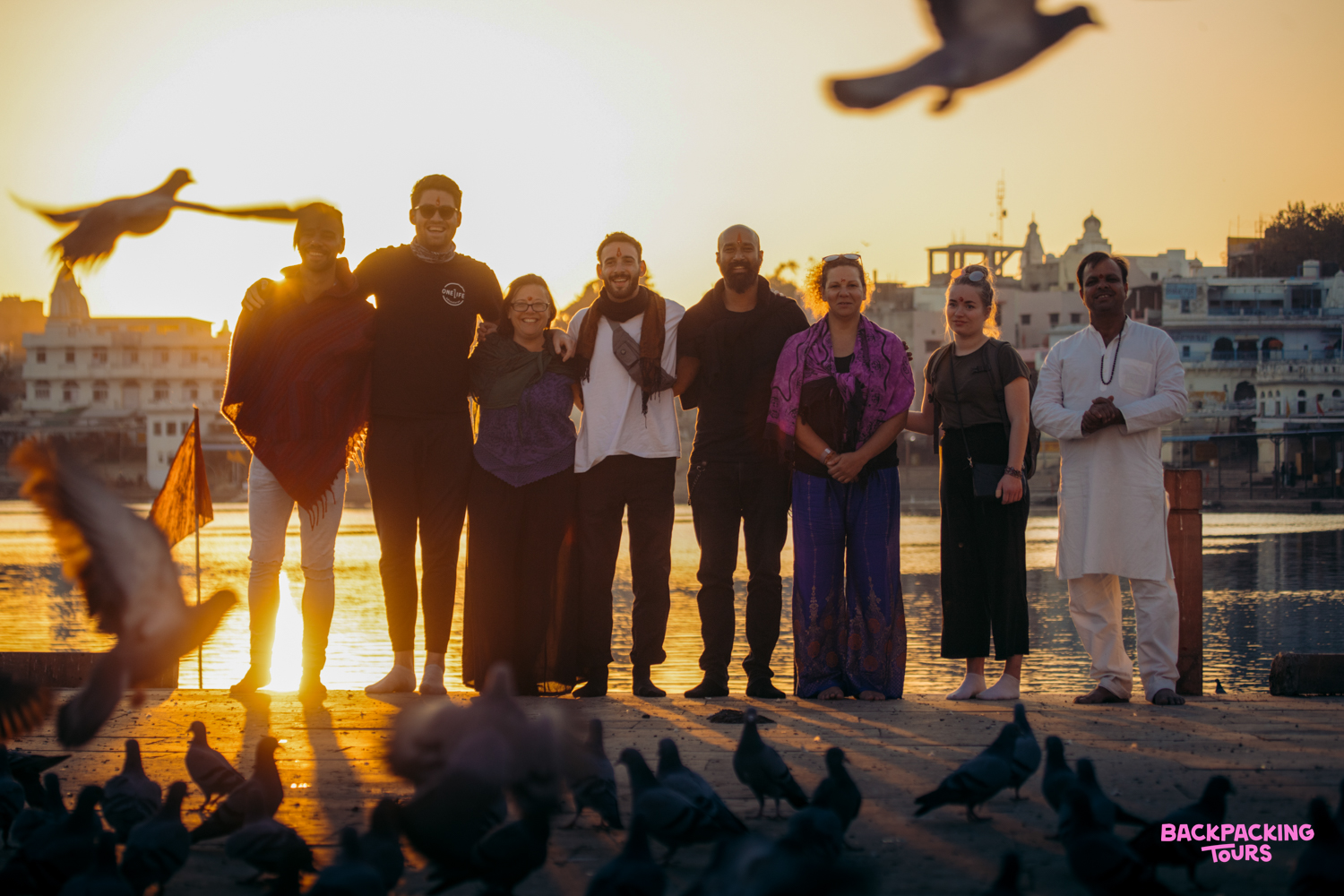 A group of travelers and a local stand by Pushkar Lake at sunset, with pigeons in the foreground.