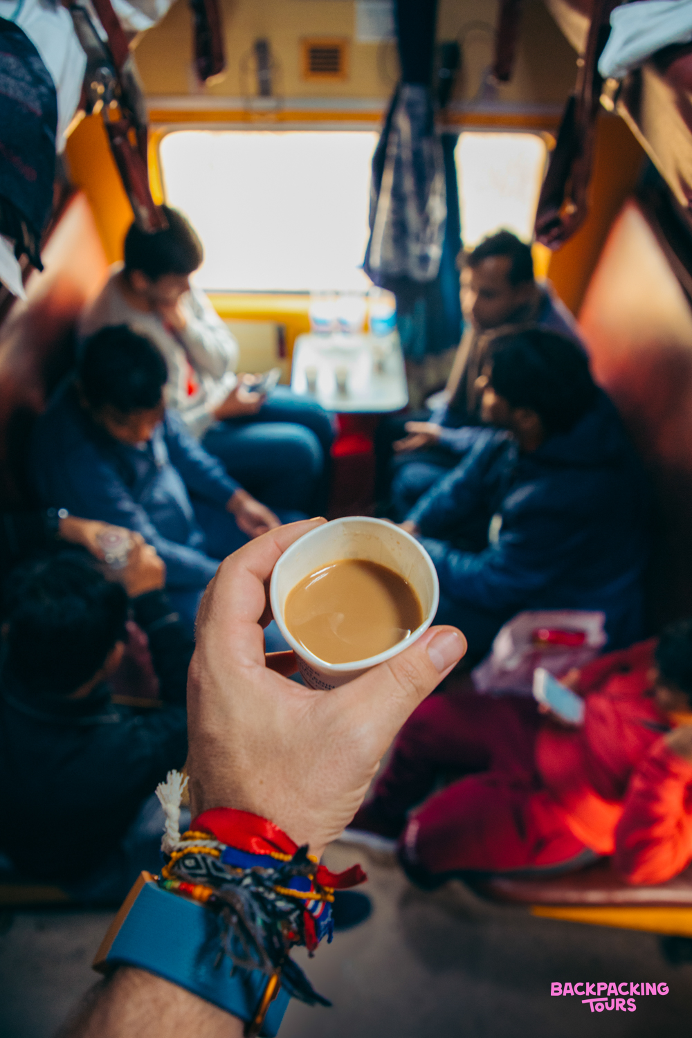 A traveler holds a cup of chai on an Indian train, with fellow passengers relaxing in the background.