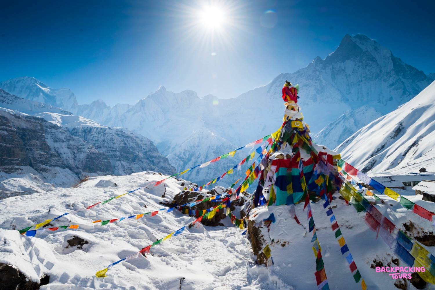 Colorful prayer flags adorn a snow-covered cairn with majestic Annapurna mountains under a bright sun.