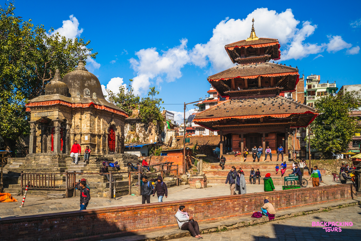People gather around ancient temples in Kathmandu Durbar Square under a clear blue sky.