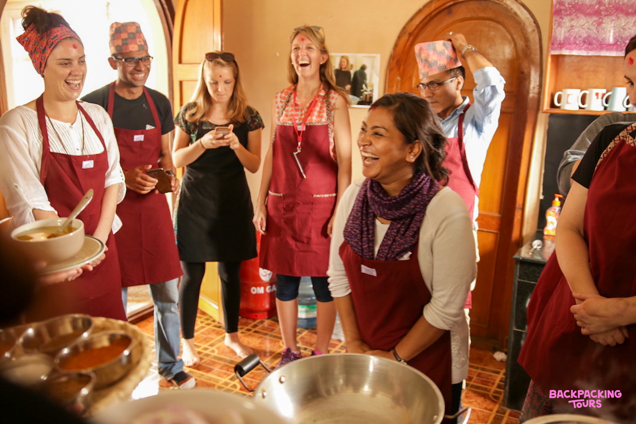 Smiling travelers and local women enjoying a lively cooking demonstration in Kathmandu, Nepal.