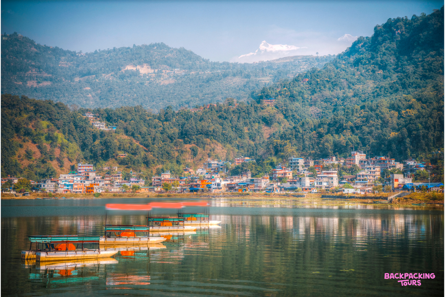 Boats float on Phewa Lake with a vibrant town and majestic Himalayan mountains in Pokhara, Nepal.