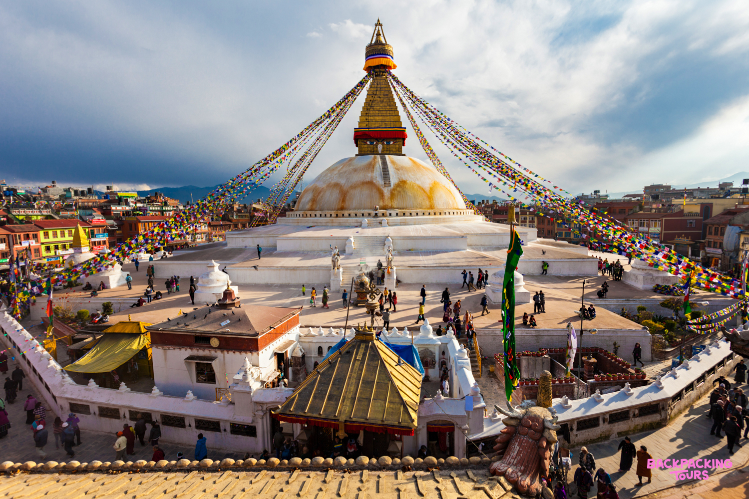Vibrant prayer flags adorn the majestic Swoyambhunath Stupa, bustling with people under a partly cloudy sky in Kathmandu.