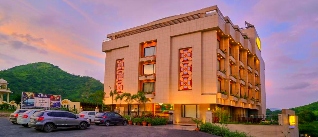 A modern hotel building with illuminated windows and cars parked outside in Udaipur at sunset.