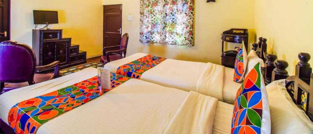 A bright hotel room featuring two single beds with colorful patterned runners and pillows.