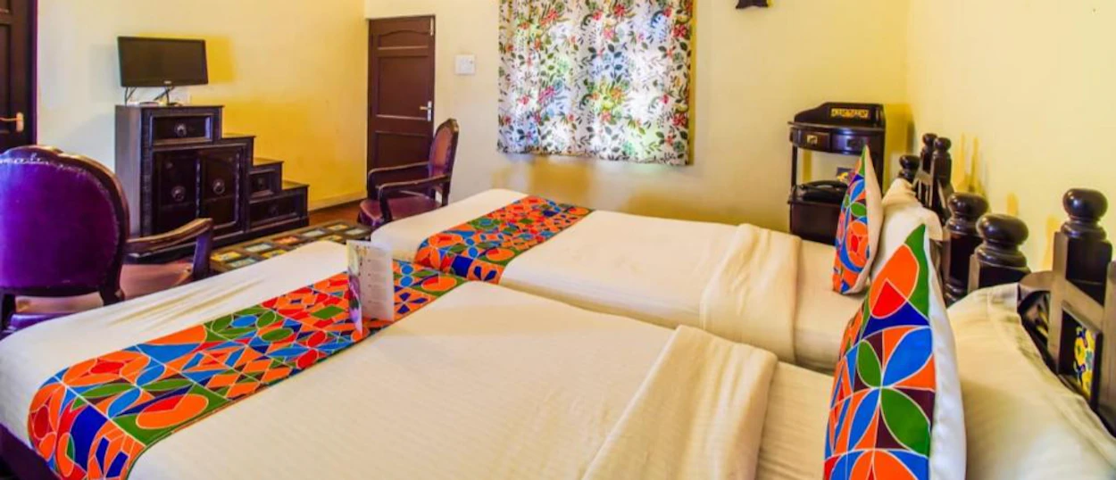 A bright hotel room featuring two single beds with colorful patterned runners and pillows.