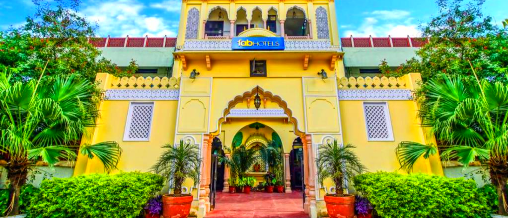 A vibrant yellow hotel building with traditional Indian architecture and lush green plants in Jaipur.