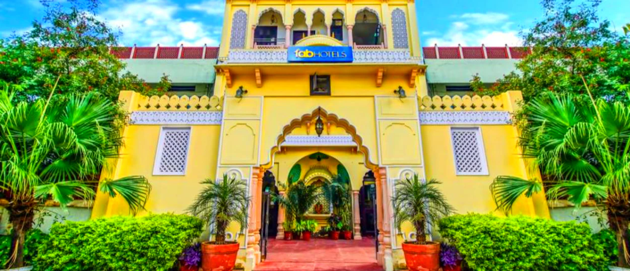 A vibrant yellow hotel building with traditional Indian architecture and lush green plants in Jaipur.