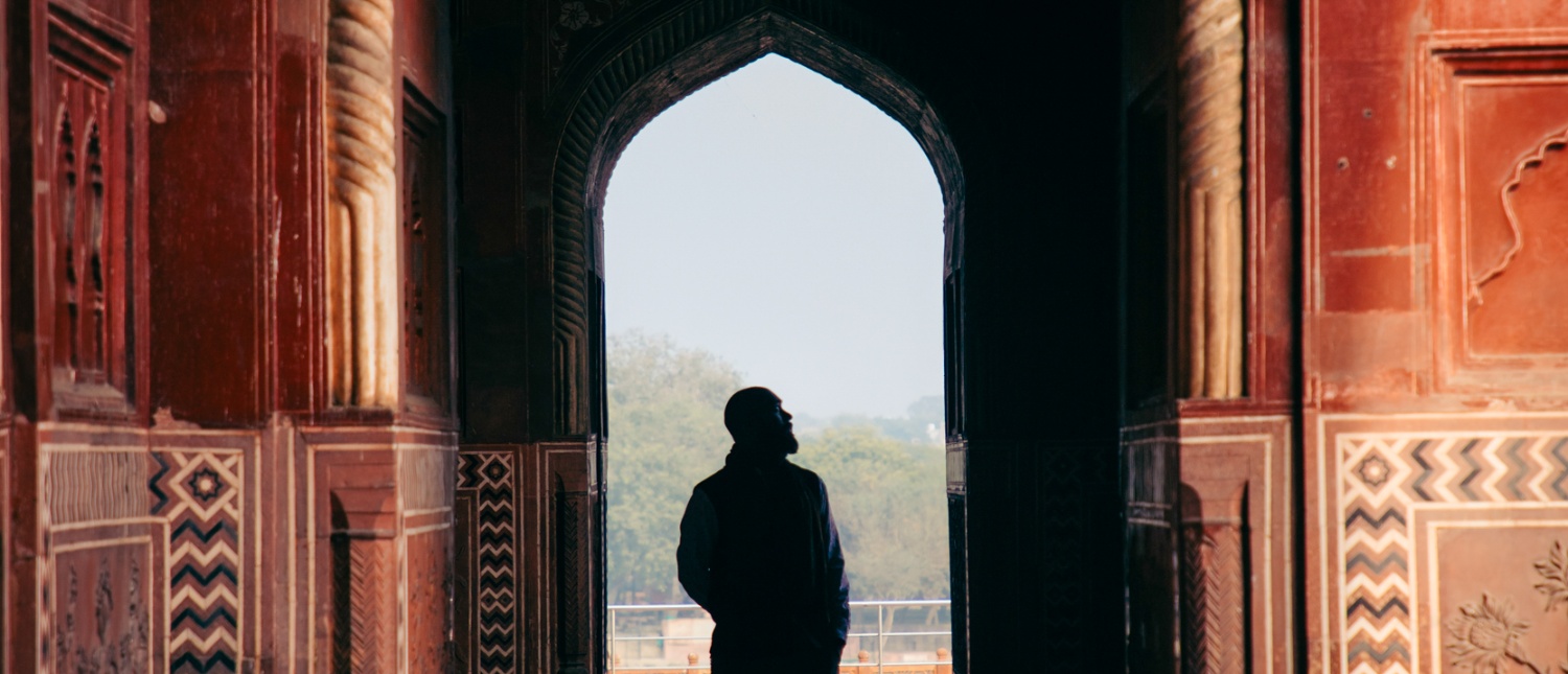 A person stands silhouetted in a decorative red stone archway at Agra Fort, looking out at a hazy sky.