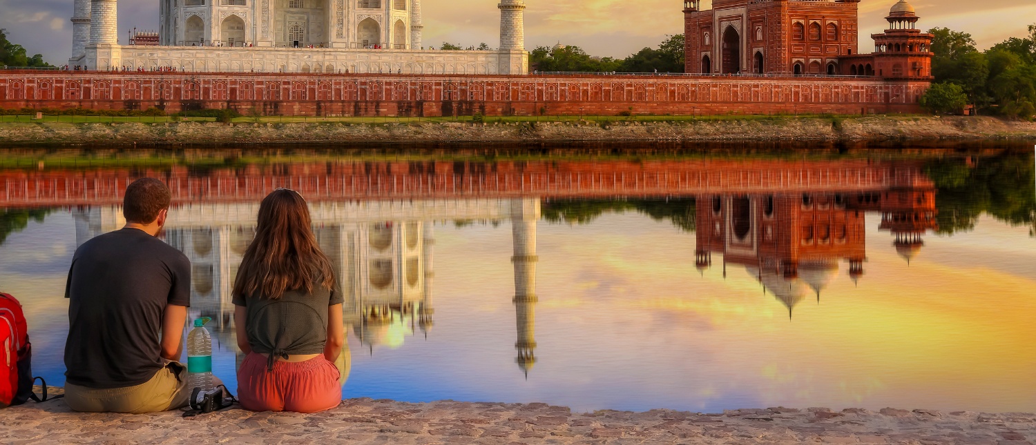 Two travelers sit peacefully, gazing at the iconic Taj Mahal and its stunning reflection in Agra.
