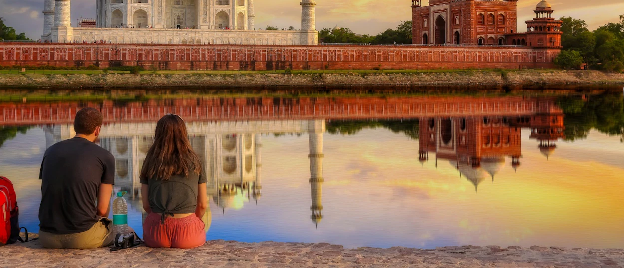 Two travelers sit peacefully, gazing at the iconic Taj Mahal and its stunning reflection in Agra.