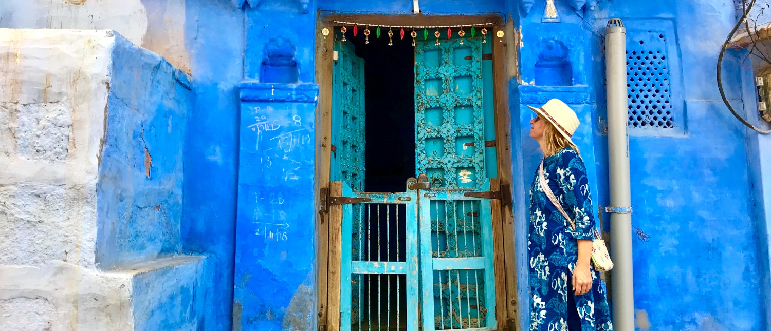A woman in a patterned blue dress and hat stands by a vibrant blue door in Jodhpur.