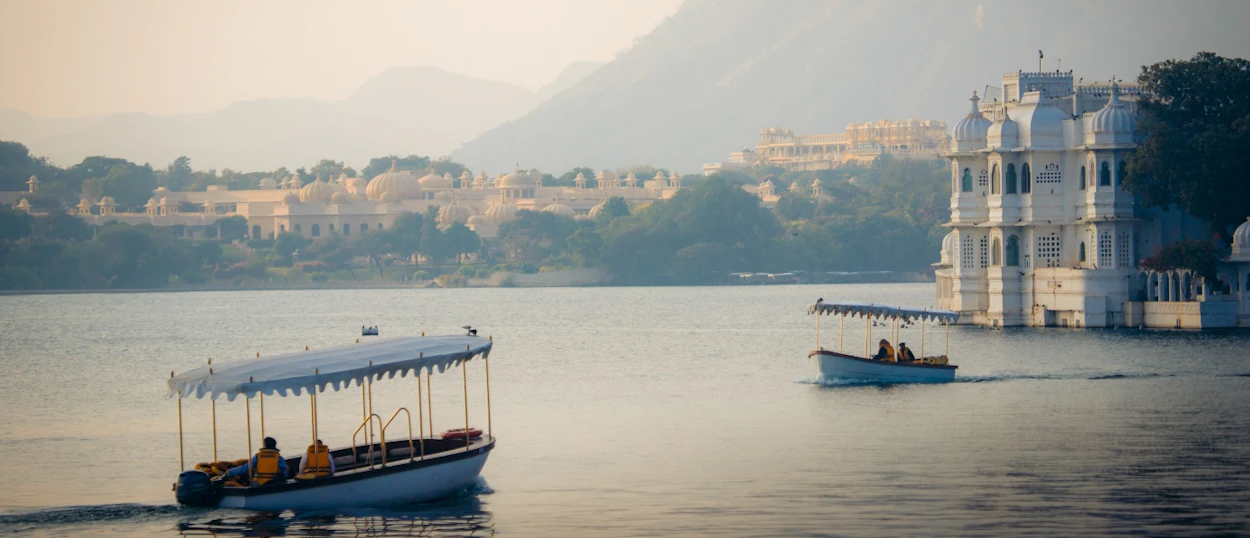 Boats with people cruise on Lake Pichola, passing white palaces and distant mountains in Udaipur.