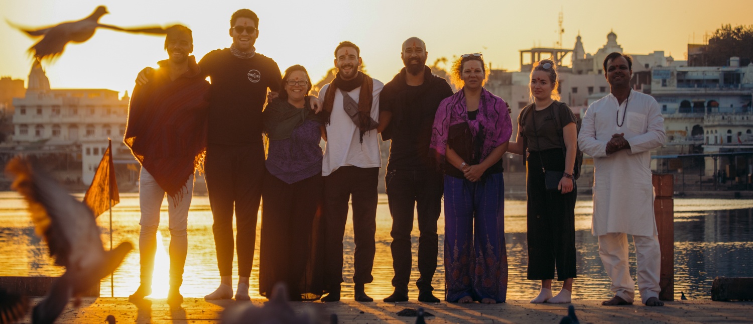 A diverse group of travelers and a local stand by Pushkar Lake at sunset with birds flying.