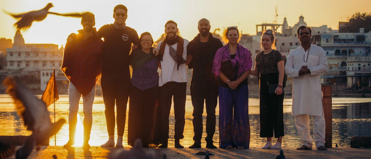 A diverse group of travelers and a local stand by Pushkar Lake at sunset with birds flying.