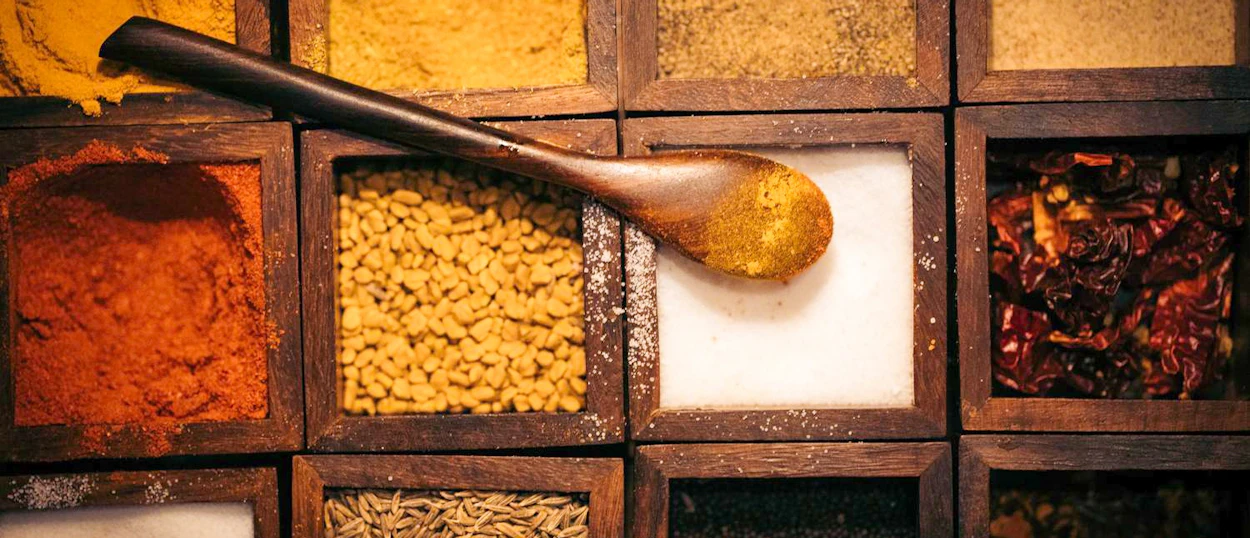 A wooden spoon filled with yellow spice rests on a vibrant box of North Indian cooking spices.