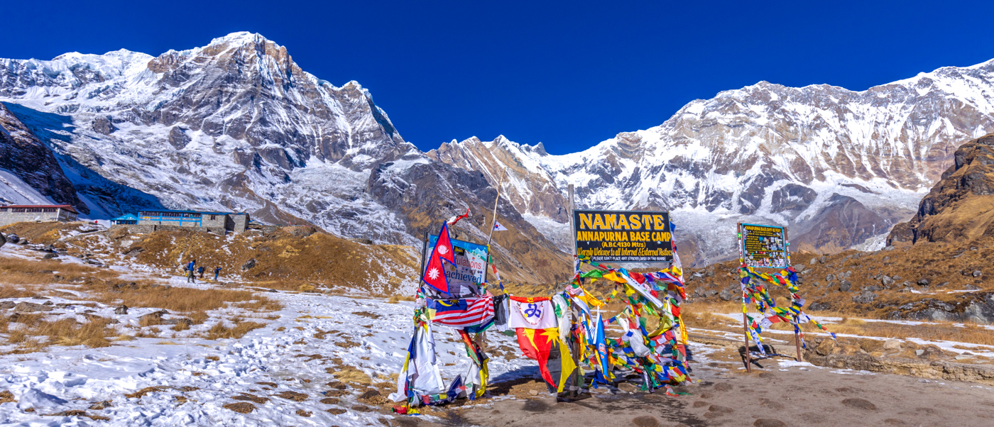 Colorful prayer flags mark the snowy Annapurna Base Camp with majestic mountains under a clear blue sky.