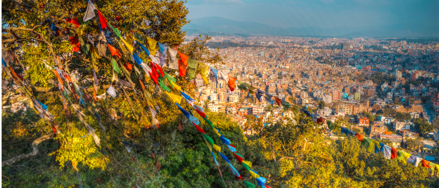 Colorful prayer flags wave over a sprawling city view of Kathmandu at sunset.